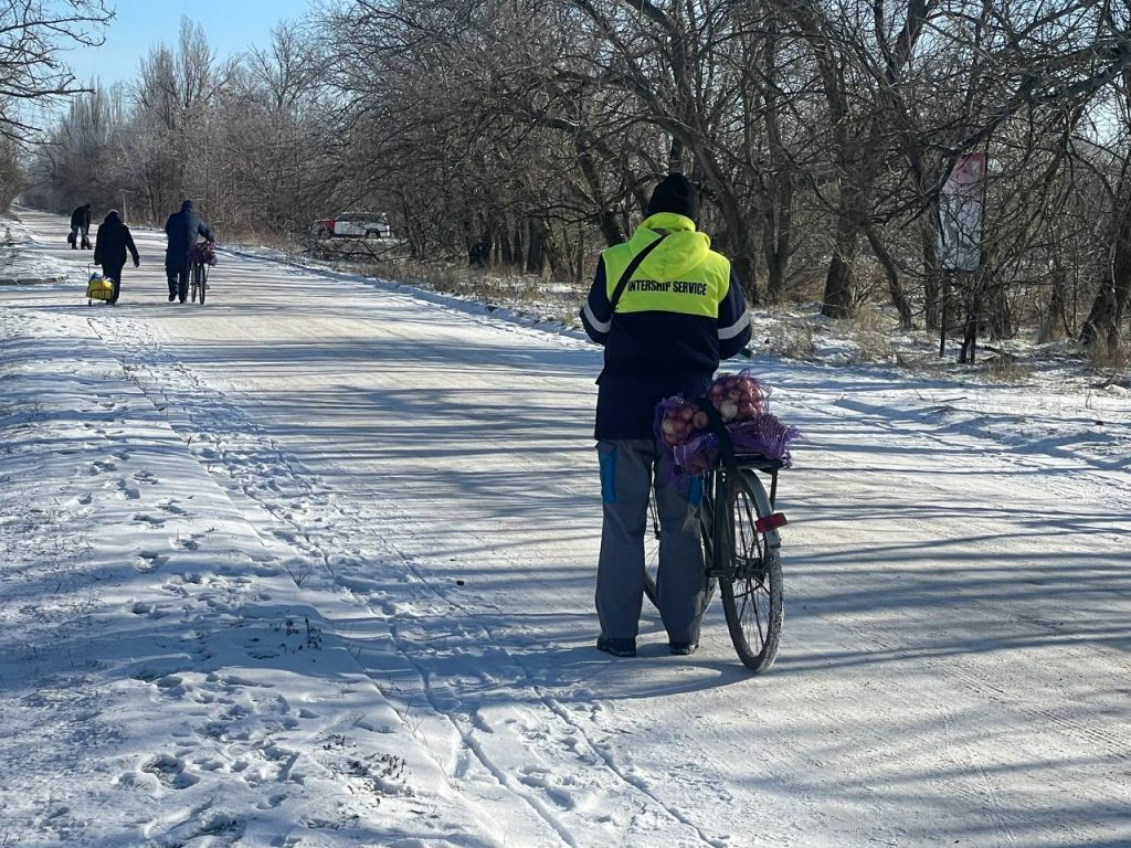 Людина везе овочі велосипедом уздовж засніженої сільської дороги